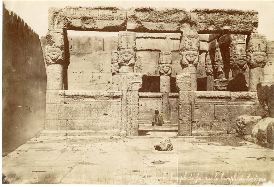 Fotografia della cappella dell'irradiazione solare (o chiosco solare) sul tetto del tempio di Hathor a Dendera, con egiziano seduto che guarda all’obiettivo. Da notare la presenza di capitelli hathorici, ancora in cima alle colonne. In basso a sinistra si trova la firma (e possibilmente la data) dell’autore.