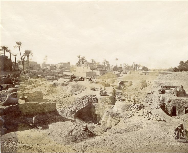 The photograph shows a number of potters at work in the open-air workshop in a suburb on the western shore near Qena, in an area close to the Temple of Hathor at Dendera, part of which is identifiable in the background. The author’s signature is visble at the bottom right. 