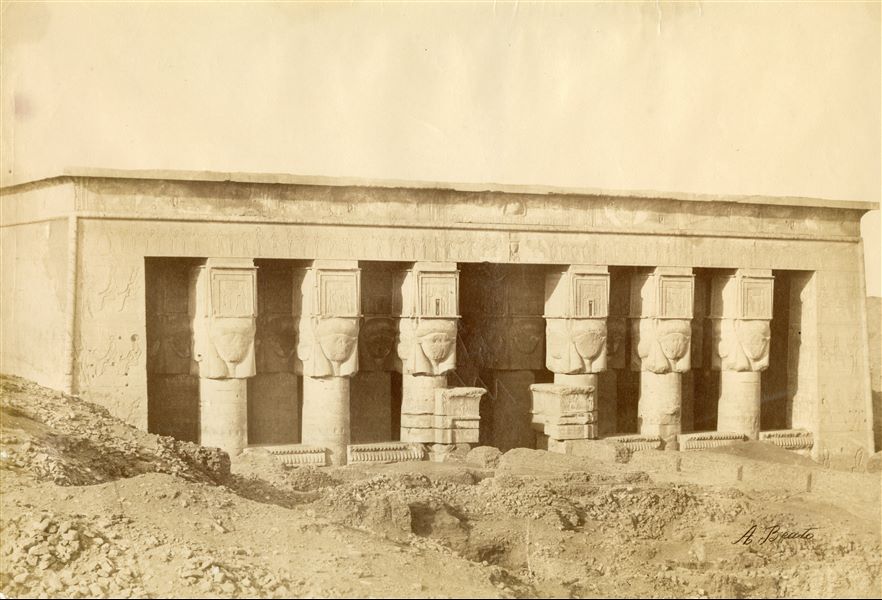 Photograph of the entrance (pronaos) to the Temple of Hathor at Dendera, showing the characteristic Hathor capitals (capitals with the head of the goddess Hathor carved in relief on each side of the columns), as well as the mud-brick structures in front of it. The author's signature can be found at the bottom right. 