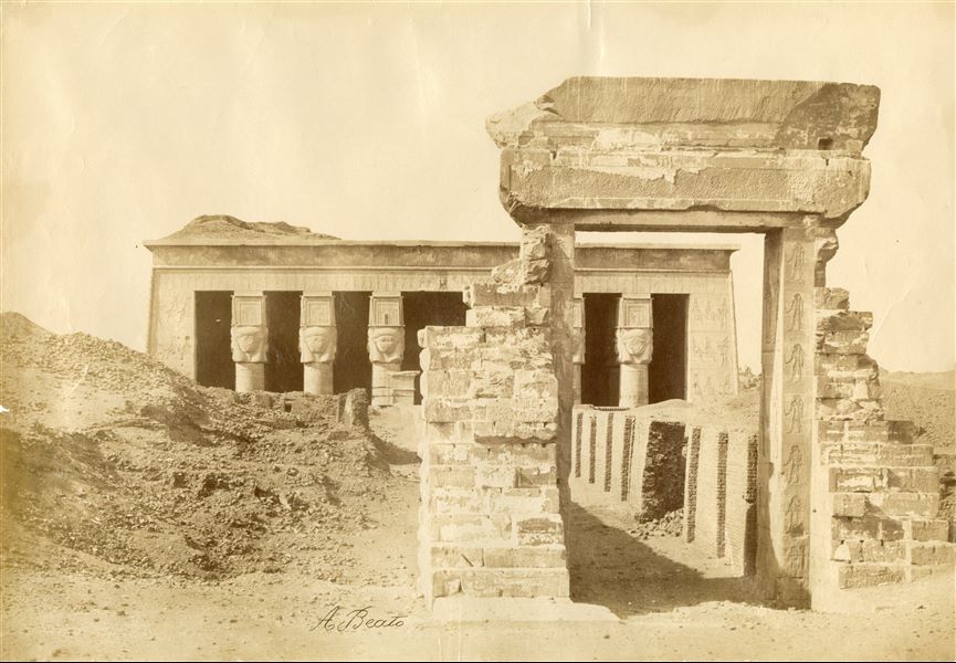 Nella fotografia si vedono il portale d'accesso e il pronao con colonne hathoriche del Tempio di Dendera. La firma dell'autore è un basso.
