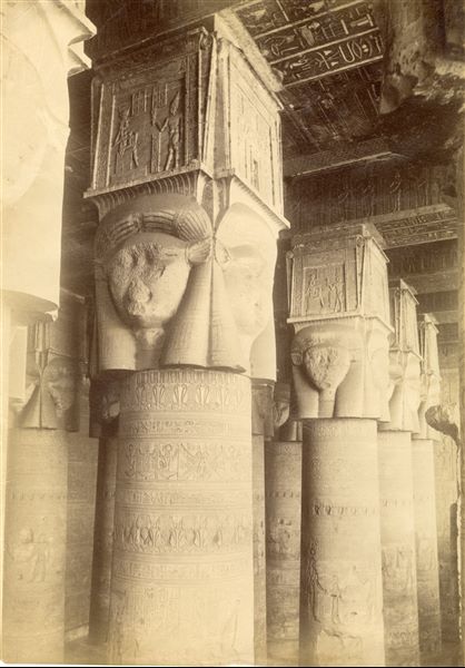 The photograph depicts the colonnade of the Hypostyle Hall, dating to the Roman Period,  from the Temple of Hathor at Dendera. Note the inscriptions on the ceiling and the Hathor capitals (capitals with the head of the goddess Hathor carved in relief on each side of the columns) further decorated with sacred scenes. The shot can be attributed to Antonio Beato. 