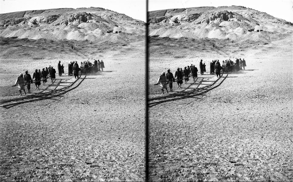 Transporting heavy loads with "Decauville" railway tracks. Some workers push the load on the trolley while others work ahead of the group by positioning the next section of tracks to create a pathway. Schiaparelli excavations. 