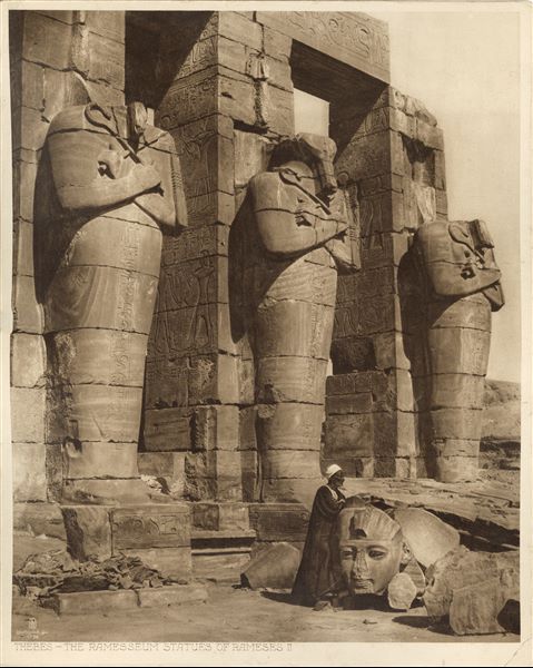 The photograph shows three Osiride pillars of Ramesses II in his Funerary Temple (the Ramesseum) in West Thebes. An Egyptian poses leaning against a fragmentary head of a fallen colossal statue.  