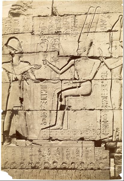 The photograph shows a detail from the interior wall decoration in the Ramesseum, where Pharaoh Ramesses II (on the left) receives the emblems of power from the god Amun seated in the centre. Next to Amun is the goddess Mut. The lower part of the photograph is blurred.