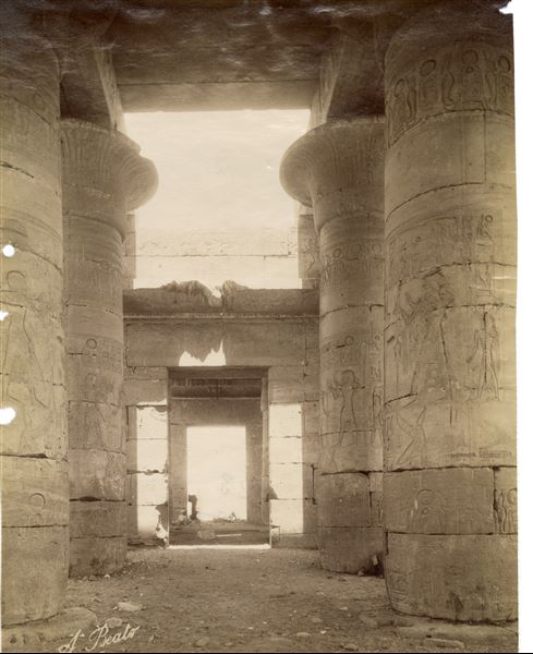 The photograph shows the central corridor of the Hypostyle Hall in the Ramesseum, the Funerary Temple of Pharaoh Ramesses II built on the west bank of the Nile at Thebes. The four end columns and the door on the western wall of the hall can be seen, near which two local inhabitants stand. The author's signature is visible at the bottom.