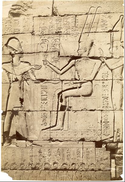 La fotografia presenta un dettaglio della decorazione parietale interna del Ramesseo, in cui il faraone Ramesse II, a sinistra, riceve gli emblemi del potere da parte del dio Amon seduto al centro. Accanto ad Amon, la dea Mut. La parte inferiore della fotografia risulta sfocata. 