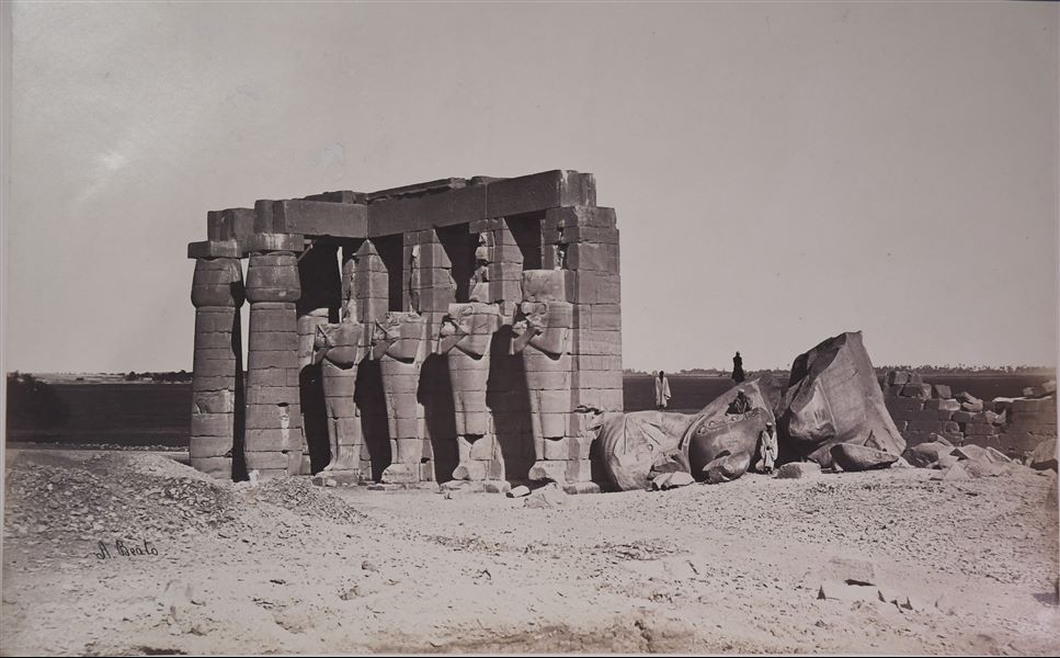 Detail of a section from the Ramesseum, the Funerary Temple of Ramesses II, where four Osiride pillars can be seen, almost all of them missing their heads. On one of the statue fragments lying on the ground, there is a graffito with the date 1870, which allows the photograph to be dated to a later period. The author's signature is visible at the bottom left. 