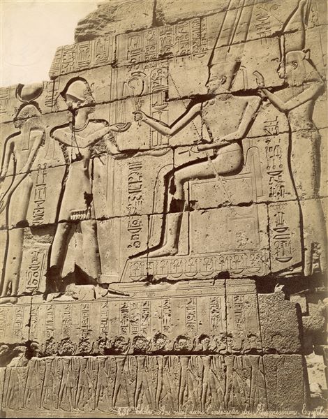 The photograph shows a detail from the interior wall decoration in the Ramesseum, where Pharaoh Ramesses II, (on the left) receives the emblems of power from the god Amun who is seated in the centre. Next to Amun is the goddess Mut. The author's signature, written with very small lettering is visible at the bottom right.