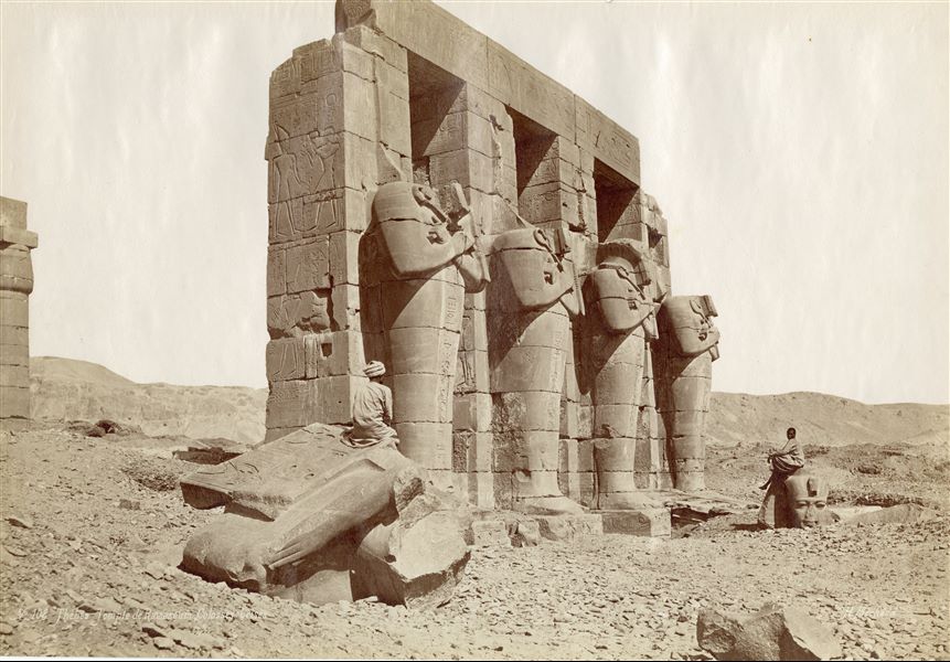 The photograph shows four Osiride pillars of Ramesses II in his Funerary Temple (the Ramesseum) in West Thebes. Two Egyptians pose seated on the colossal fragments of some Ramesside statues. The author has placed his signature at the bottom right. 