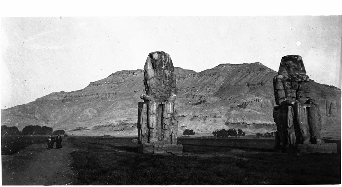 View of the so-called Colossi of Memnon. A pair of colossal statues depicting the pharaoh Amenhotep III seated on a throne. The Theban mountain is visible in the background. 