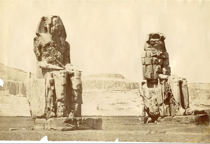 The photograph depicts the so-called Colossi of Memnon (in the foreground) in West Thebes, two enormous statues commissioned by Pharaoh Amenhotep III placed in front of his funerary temple. Some egyptians are sitting on the base of the statue on the left. Part of the Theban mountain is in the background, with some of the tombs of nobles visible. The author's signature is visible at the bottom. 