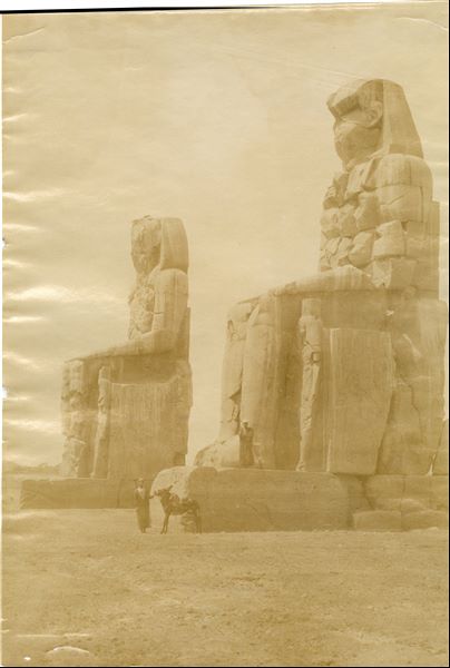 The photograph shows the so-called Colossi of Memnon (in the foreground) in West Thebes. In the background, on the right, the remains and Osiride pillars of the Ramesseum (the Funerary Temple of Ramesses II) are clearly visible. 