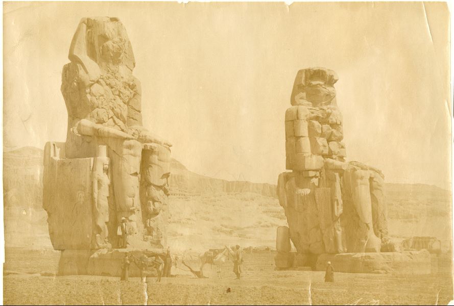 In the foreground the Colossi of Memnon, with egyptians and two donkeys nearby. The Theban mountain, is in the background, with temples (the ruins of the Ramesseum can be glimpsed on the right) and rock-cut tombs. The author's signature is visible at the bottom right. 
