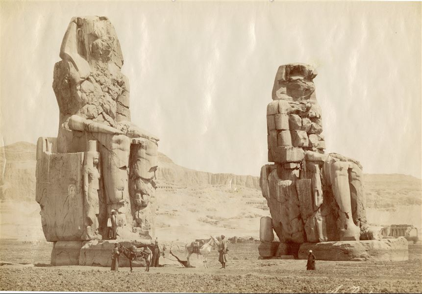 In the foreground the Colossi of Memnon, with egyptians and two donkeys nearby. The Theban mountain, is in the background, with temples (the ruins of the Ramesseum can be glimpsed on the right) and rock-cut tombs. The author's signature is visible at the bottom right.  
