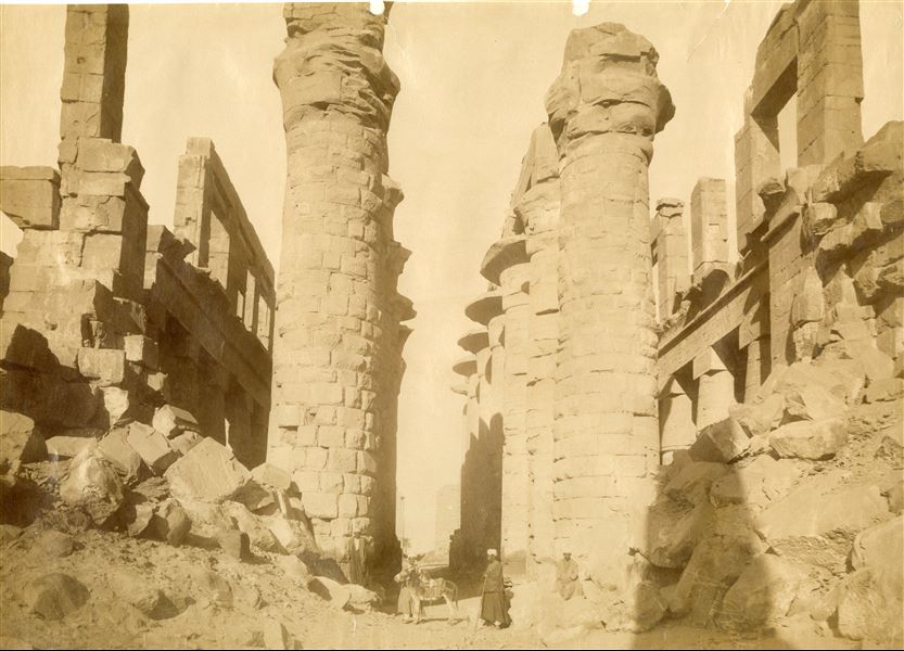 The photograph shows the central colonnade of the Great Hypostyle Hall in the Karnak Temple Complex, in the direction of the second and first pylons, with four egyptians and a donkey at the foot of the ruins. Note the accumulation of blocks on either side of the entrance. The author's signature can be found at the bottom right.  