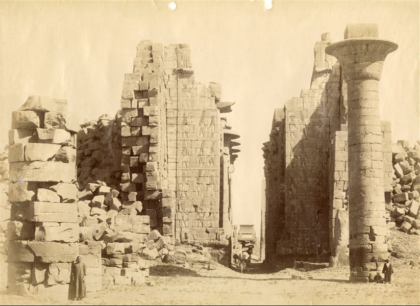 The photograph depicts the entrance to the Great Hypostyle Hall, built by Pharaoh Seti I, dof the Karnak Temple complex, viewed from the courtyard in front. The remaining column from the south-eastern corner of the kiosk of Taharqa (ruler of the 25th dynasty) stands out. Some locals (seated and standing) and a donkey give an idea of the colossal size of the complex. The author's signature is visible at the bottom centre.   