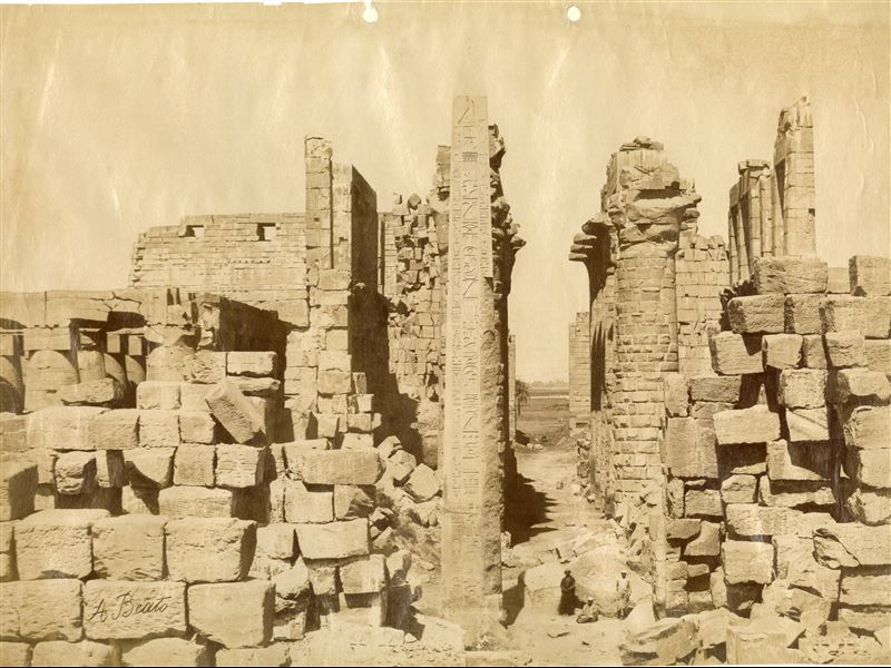 La fotografia presenta una veduta delle rovine del complesso templare di Amon a Karnak, con al centro l’obelisco del faraone Thutmosi I, della XVIII Dinastia, e tre abitanti locali in posa accanto al monumento, per dare un'idea delle sue dimensioni. Sullo sfondo, la Grande Sala Ipostila. In basso si trova la firma dell’autore.  