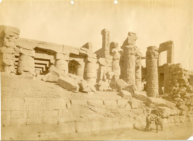View of the south-western, external wall of the Great Hypostyle Hall built by Seti I in the Karnak Temple Complex, with one figure in the foreground and another at the height of the colonnade (in the background on the right). The ground level still covers part of the wall. The author's signature (partially cut off) is legible at the bottom. 