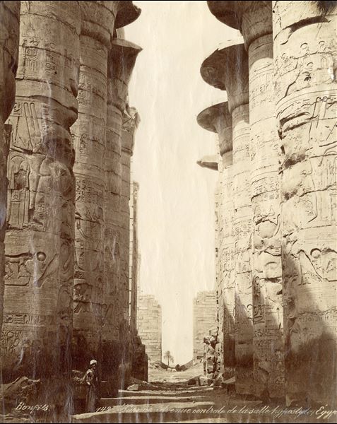 The image shows a view of the central corridor of the Great Hypostyle Hall, built by Pharaoh Seti I, in the Karnak Temple Complex. A man in Egyptian clothing is looking towards the camera. The author's signature can be found at the bottom left. 