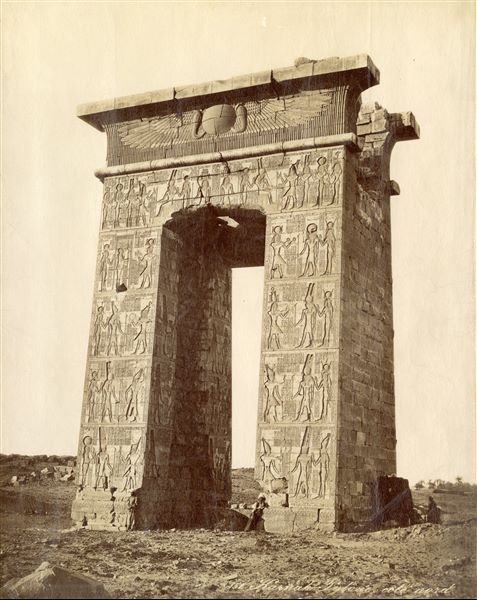The photograph shows the entrance gate, on the southern side, built by Ptolemy III Euergetes in the Karnak Temple Complex. Some egyptians are photographed next to the darkroom, the black curtain visible next to the gate was perhaps used for developing film. Based on the style of the handwriting, the work can be attributed to F. Bonfils. 