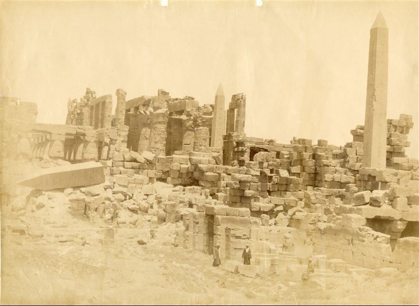 The image shows the ruins of the Karnak Temple Complex, in particular the 18th dynasty buildings (right) and the Ramesside Great Hypostyle Hall (left). Among the ruins stand the obelisks of Hatshepsut (north, right) and Tuthmosis I (south, centre); note that the tip of the collapsed second obelisk of Queen Hatshepsut. Two egyptians look towards the camera lens. The author's almost completely faded signature is at the bottom left.   