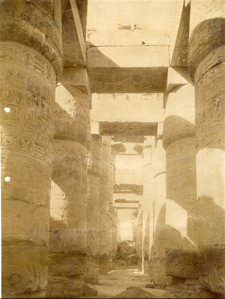 The photograph shows an interior view of the Great Hypostyle Hall of the Karnak Temple Complex, where the distinctive "Window of Appearance" can be seen at the top. Note that the ground level still covers the columns' bases. In the background are some debris and blocks that have not yet been put back in place.   