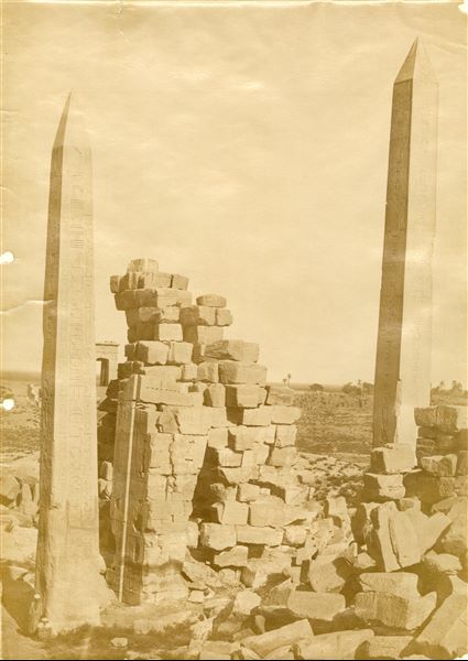 The photograph shows the 18th dynasty obelisks in the Karnak Temple Complex seen from the south. The obelisk of Tuthmosis I is on the left and that of Hatshepsut is on the right. Two people, at the foot of the older monument, allow the photographer to demonstrate the size of the monuments. In the background, the Gateway of Ptolemy III is visible.  