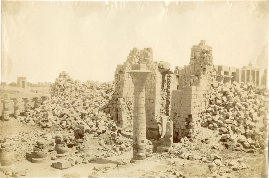 The photograph shows the collapsed second pylon in the first courtyard of the Karnak Temple Complex, where the Great Hypostyle Hall from the Ramesside period was built behind it. In the foreground, the last surviving column of the kiosk erected by Pharaoh Taharqa is visible. Behind the hall, in the background, the tips of the two obelisks still standing on site, those of Tuthmosis I and Hatshepsut can be seen.  