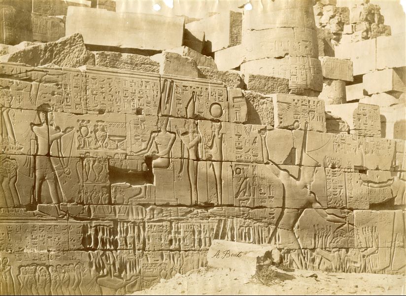 Veduta di una porzione della parete settentrionale esterna della Grande Sala Ipostila del complesso templare di Amon a Karnak. Il faraone Seti I è rappresentato durante le campagne militari a destra) e mentre dedica la vittoria sui nemici ad Amon (in alto a sinistra). In basso la firma dell’autore.