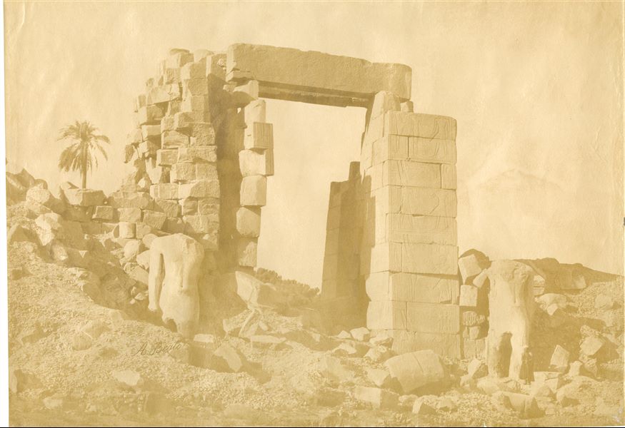 The photograph shows the northern side of the yet to be restored tenth pylon in the Karnak Temple Complex, built by Pharaoh Horemheb, with two colossal statues of the pharaoh (later reused by Ramesses II) on either side of the gate. The ground level still covers the lower part of the colossal statues. The author's signature is visible at the bottom left. 