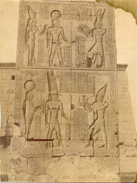 La fotografia presenta un dettaglio delle iscrizioni sul portale meridionale di accesso al complesso templare di Amon a Karnak, fatta costruire dal faraone Tolemeo III Evergete. Dietro al monumento, si scorge il lato orientale della facciata del tempio di Khonsu. In basso a destra, la firma dell’autore.