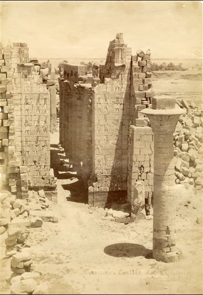 La fotografia mostra il primo cortile del complesso templare di Amon a Karnak, dietro cui si erge il II pilone e la Grande Sala Ipostila, con i resti del chiosco del faraone Taharqa a destra (rappresentato dalla colonna ancora stante), visti dal I pilone, da cui si accede al complesso. In basso, la firma dell’autore. Fotografia scattata probabilmente dalla sommità del I pilone.