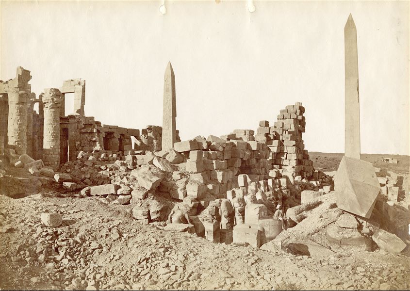 La fotografia mostra una veduta da sud della Sala Wadjet del complesso templare di Amon a Karnak, costruita dal faraone Thutmosi I, oltre che i due obelischi di Hatshepsut (uno stante, l’altro crollato). Sullo sfondo, al centro, si riconosce anche l'obelisco superstite di Thutmosi I. Un egiziano siede su uno dei basamenti delle imponenti colonne. In basso a destra, la firma dell’autore.  