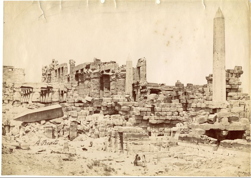 The photograph shows an eastern  view of the colonnaded Wadjet Hall built by Pharaoh Tuthmosis I in the Karnak Temple Complex. The still standing obelisks of Hatshepsut (north, on the right), the tip of he collapsed one (south, on the left) and that of Tuthmosis I (centre) are also visible. In the background, the western ruins of the Great Hypostyle Hall of Seti I can be seen. Two egyptians seated on a boulder in front of the temple, pose for the photographer, whose signature is legible on the left. 
