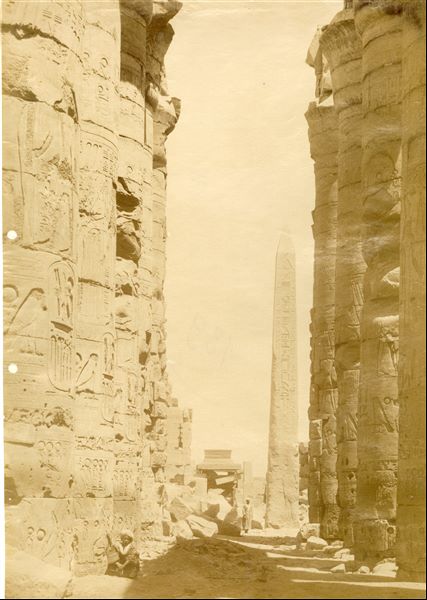 The picture shows a view of the central corridor of the Great Hypostyle Hall commissioned by Pharaoh Seti I in the Karnak Temple Complex, ending with the third pylon. In the forecourt between the third and fourth pylons, the surviving obelisk of Pharaoh Tuthmosis I is visible. The photo can be attributed to Beato.