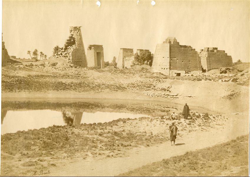 The photograph depicts the ruins of two pylons in the Karnak Temple Complex, seen from the Sacred Lake, specifically the ninth pylon (left, that of Pharaoh Horemheb) and the eighth pylon (right, that of Queen Hatshepsut). In the background, centre, the southern gate commissioned by Ptolemy III Euergetes to access to the Temple of Khonsu, from where the pylon is visible. The perspective however, is distorted but the two monuments are aligned. Three egyptians on the bank of the slightly drained Sacred Lake look towards the camera lens. The author's signature is visible at the bottom left.  
