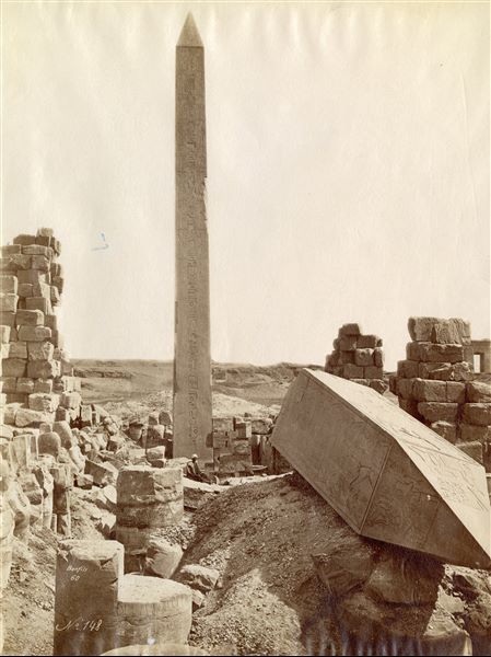 The picture captures the two obelisks of Hatshepsut near the Wadjet Hall of Pharaoh Tuthmosis I in the Temple of Amun at Karnak, one obelisk is still intact, whilst the other (in the foreground) has its tip on the ground. An egyptian sits at the base of the monument in the background. A signature and presumably a date are visible at the lower left.  