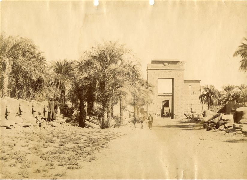 The image shows the Avenue of Sphinxes with ram's heads, leading to the southern entrance, built by Ptolemy III Euergetes, in the Karnak Temple Complex. In the background, the entrance to the Temple of Khonsu is visible, aligned with the entrance. Some egyptians look towards the camera lens, while others continue their work. 