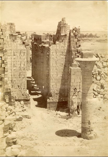 The photograph shows the first courtyard of the Karnak Temple Complex, with the second pylon behind it and the Great Hypostyle Hall, with the remains of the kiosk of the Pharaoh Taharqa on the right (represented by the column still standing). All viewed from the first pylon, from which the complex is entered. The author's signature is visible at the bottom. Photograph probably taken from the top of the I pylon.   