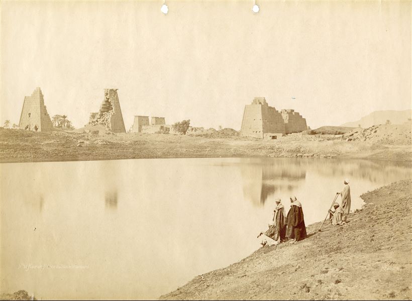 The image shows five Egyptians on the bank of the Sacred Lake of Karnak in the Karnak Temple Complex. In particular the eighth pylon (on the right, that of Queen Hatshepsut) and ninth pylon (left, that of Pharaoh Horemheb). In the background, the ruins of the Temple of Khonsu can be see within the Complex, where the pylon and the eastern outer enclosure walls are visible. The author's signature can be found at the bottom right.