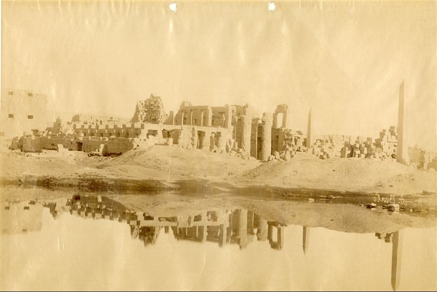 The picture shows what remains of the structural elements that make up the Ramesside Great Hypostyle Hall of the Karnak Temple Complex, behind which the first pylon can be seen. In this frame, taken from the Sacred Lake, the obelisk of Hatshepsut and that of Tuthmosis I stand out in the foreground. The author's signature which is mirrored can be found on the right.  