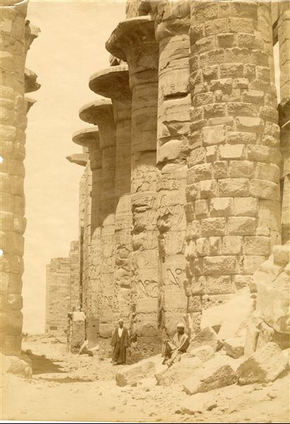 The photograph shows the central colonnade of the Great Hypostyle Hall inside the Karnak Temple Complex, with two men probably positioned to show the sheer size of the columns. In the background, to the right, part of the second pylon is visible. The shot can be attributed to Antonio Beato. 
