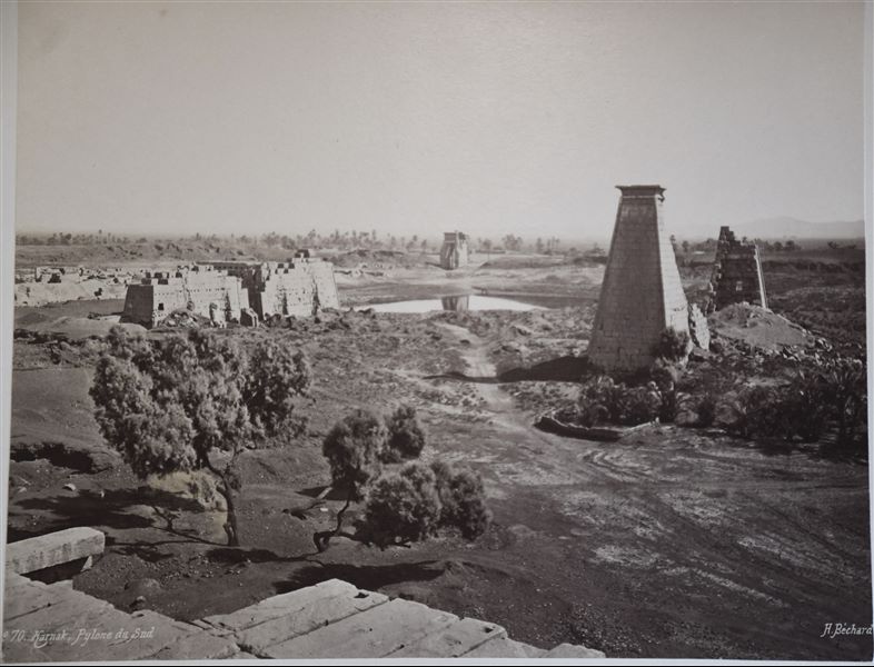 View of the Karnak Temple Complex, from the north-south axis. In particular, the remains of the seventh (far left), eighth (left) and ninth pylons (centre) are visible. Behind, the Sacred Lake, and in the background is the gate built by Nectanebo I. Photograph taken from the Temple of Khonsu. The author's signature can be found at the bottom right. 