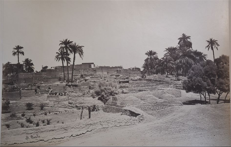 Landscape view of the village of Karnak, in Luxor. In the background the remains of the Karnak Temple Complex are visible, including an obelisk (left), and the massive structure of the Great Hypostyle Hall. The author's signature can be found at the bottom left. 