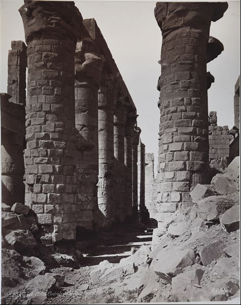 Particolare della navata centrale della Grande Sala Ipostila del complesso templare di Amon a Karnak, in direzione del II pilone. Il livello del suolo copre ancora parzialmente le basi delle colonne. La firma dell’autore si trova in basso a destra.