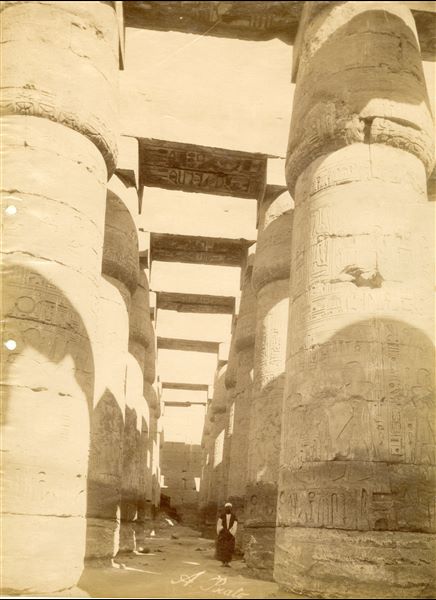 Veduta del colonnato della Grande Sala Ipostila del complesso templare di Amon a Karnak, all'ombra della quale posa un abitante locale. Da notare che il livello del terreno copre ancora le basi delle colonne. In basso al centro si trova la firma dell’autore.