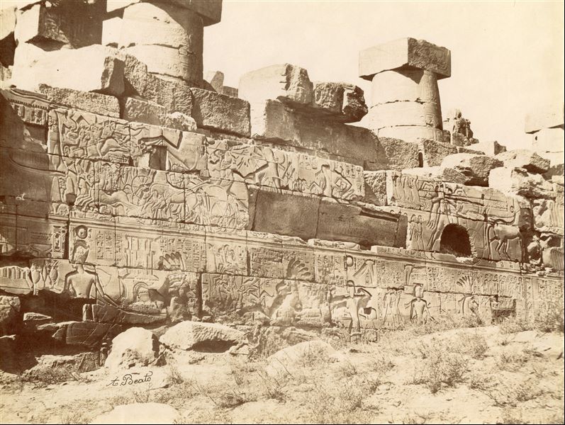 The photograph shows the bas-reliefs on the outer wall (north side, east section) of the Great Hypostyle Hall in the Karnak Temple Complex. Showing battle scenes of Pharaoh Seti I against the Yenoam  (first register from the top) and Shasu populations (second register from the top), who were slaughtered with the pharaoh's war chariot during his Syrio-Palestine campaigns. The photograph documents the ground level in the second half of the 19th century, which was a few metres higher than the original level. Part of the wall is in fact, still underground. At the bottom, the author's signature is visible. 