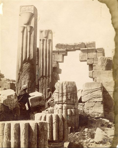 L'immagine mostra le rovine del vestibolo costruito da Thutmosi III nel complesso templare di Amon a Karnak, tra cui sono visibili i pilastri in granito rosa che recano gli emblemi dell’Alto (destra) e Basso (sinistra) Egitto, rispettivamente il loto per il Sud e il papiro per il Nord. Sopra, il cartiglio di Thutmosi III. Due egiziani sono presenti ai piedi delle colonne. In basso a destra è leggibile la firma degli autori.