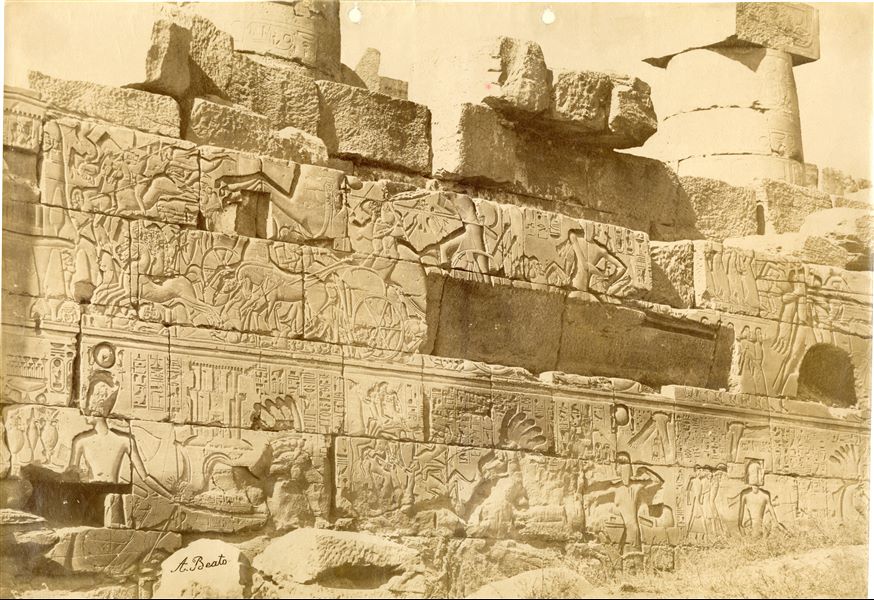 The photograph depicts a detail of the decoration of the outer wall, north side, of the Ramesside Great Hypostyle Hall in the Karnak Temple Complex. It shows Pharaoh Seti I in various stages of battle against the Syrio-Palestinians. Note that the ground level still covered part of the wall. The author's signature is clearly visible at the bottom left.  