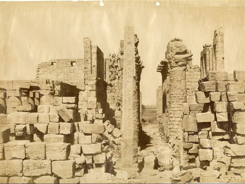 The photograph shows a view of the ruins from the Karnak Temple Complex, with the 18th dynasty obelisk of Pharaoh Tuthmosis I in the centre, and three local inhabitants posing next to the monument to give an idea of its size. In the background is the Great Hypostyle Hall. The author's signature is visible at the bottom.